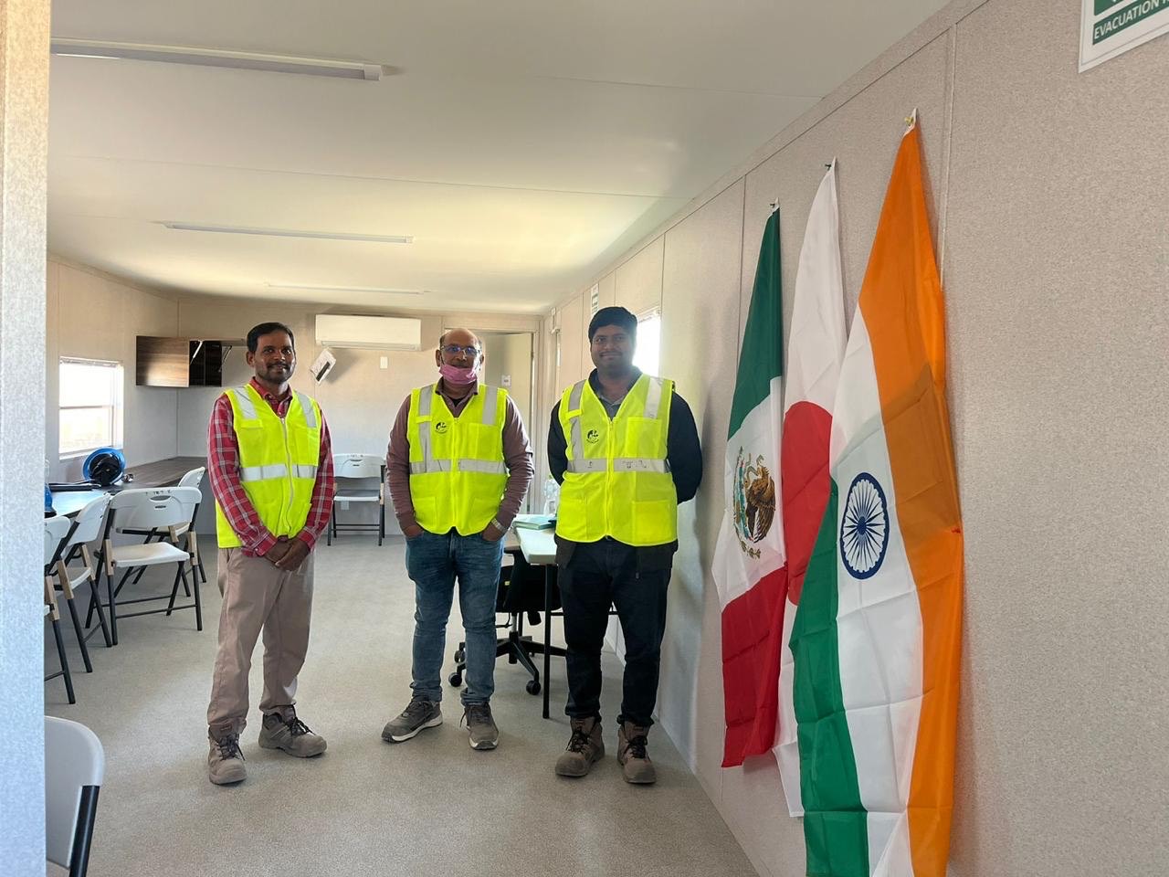 Project engineers in high-visibility vests in the site office, with Mexican and Indian flags on the wall